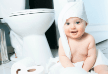 A baby wrapped in toilet paper sitting next to a toilet.