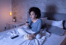 A woman alone watching TV at night drinking wine and eating popcorn.