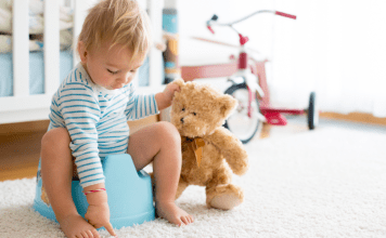 A boy sitting on a potty holding his teddy bear.