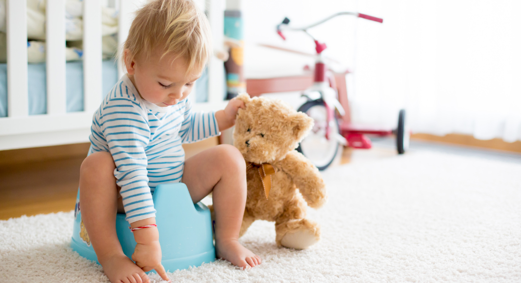 A boy sitting on a potty holding his teddy bear. 