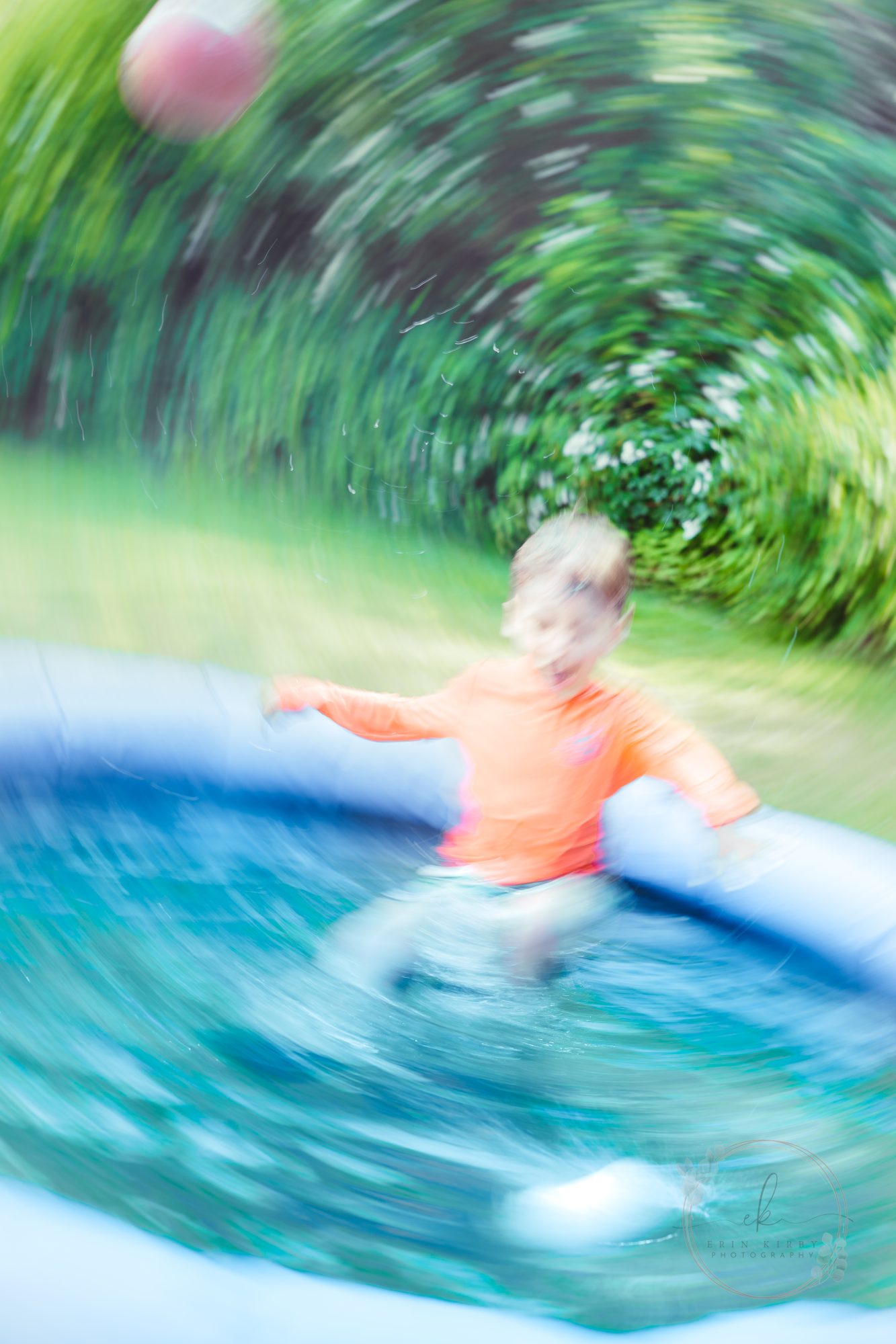 A boy with short light hair and fair skin jumps into a pool. In the background are green grass and bushes. A ball flies up in the air to the left of the screen. The image is blurred in a circle